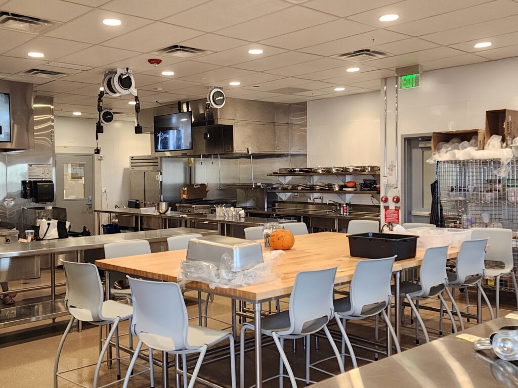 Professional commercial kitchen training area with work tables, stainless steel equipment, and seating