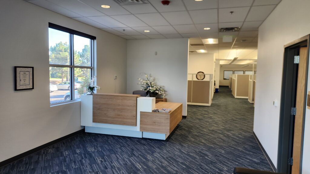 Modern office reception area with wooden desk, carpet flooring, and open workspace behind