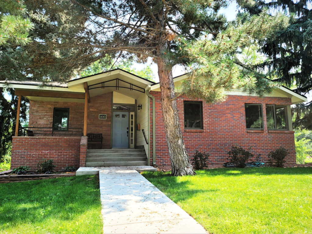 Mid-century modern brick ranch house with front porch, large trees, green lawn, and walkway entrance