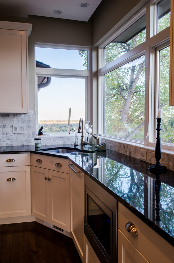 Modern kitchen featuring white cabinets, black granite countertops, stainless steel sink, and large windows overlooking trees