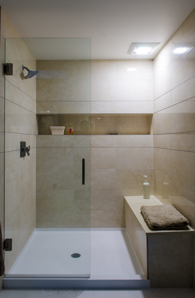 Modern bathroom walk-in shower with glass door, beige stone tiles, built-in shelf, and shower bench