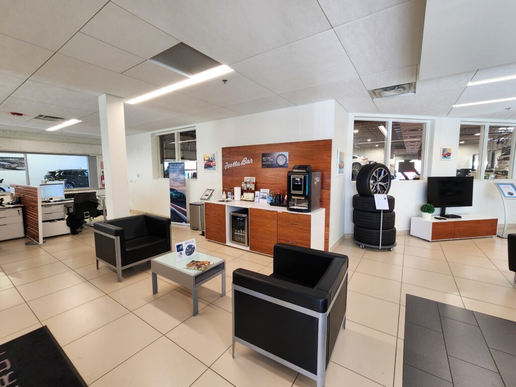 Modern BMW dealership customer lounge with coffee bar, seating area, tire display, and premium automotive showroom interior