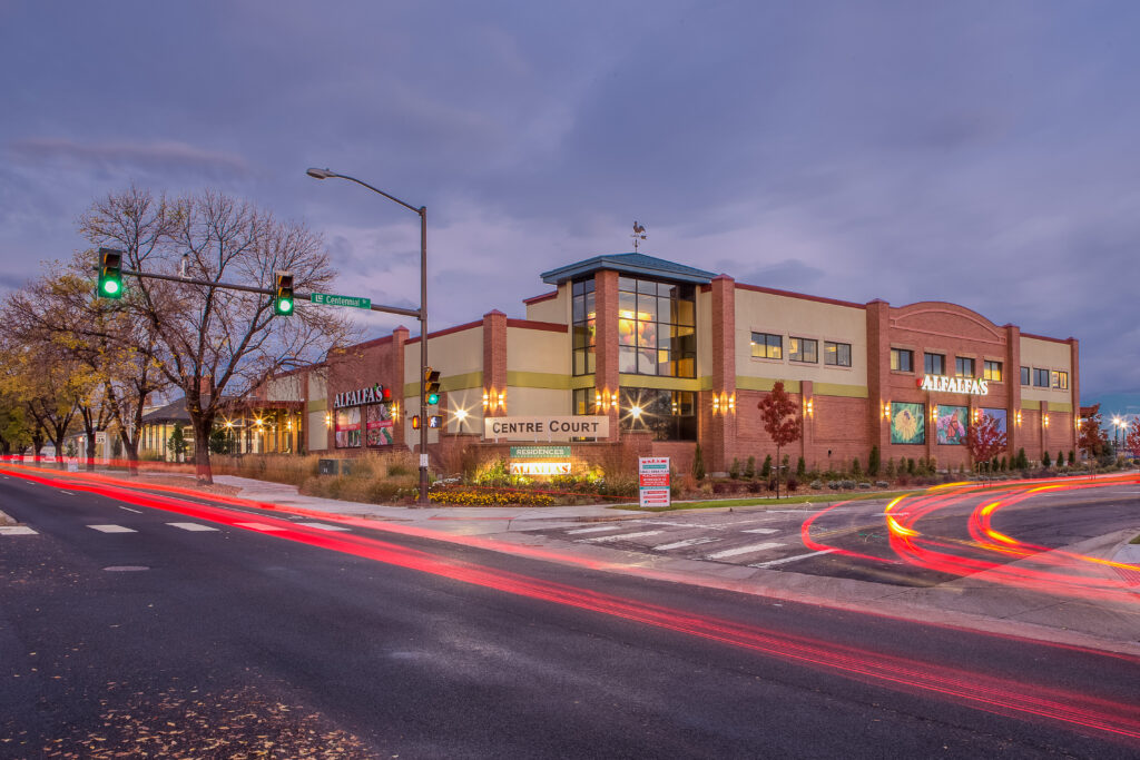 Modern commercial complex exterior with brick facade, glass entrance, landscaping, street view, and evening architectural lighting