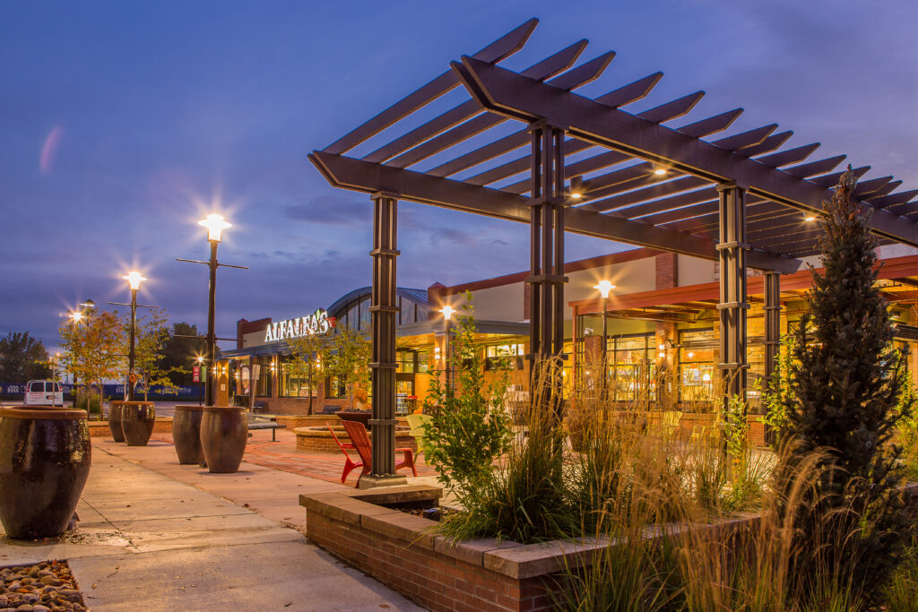 Commercial plaza exterior with modern pergola structure, landscaped walkway, outdoor seating, and illuminated building facade