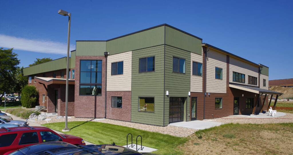 Contemporary commercial building exterior with brick facade, green metal panels, large windows, entrance canopy, and landscaped surroundings