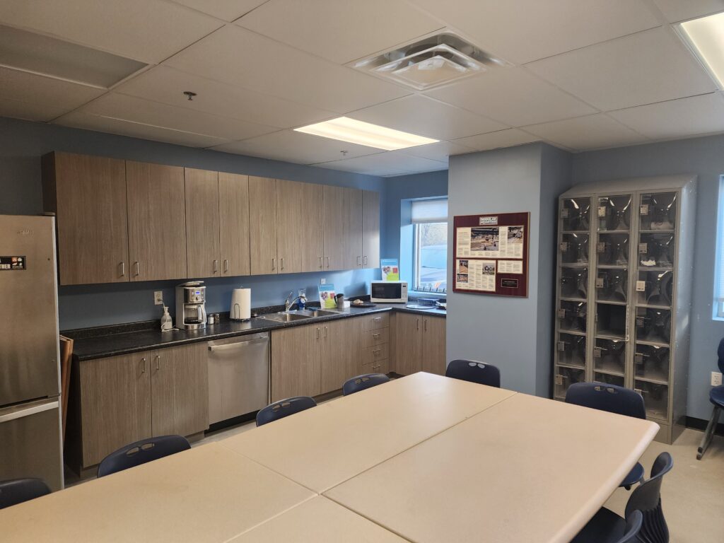 Modern industrial facility break room with kitchen cabinets, meeting table, chairs, and employee workspace area