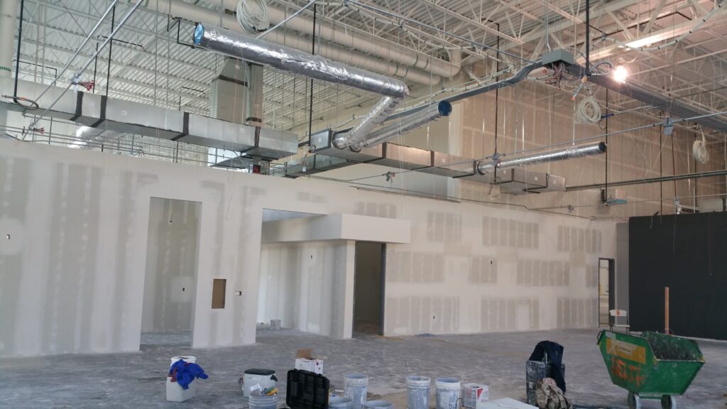 Industrial facility interior under construction with exposed HVAC ducts, electrical systems, and unfinished walls