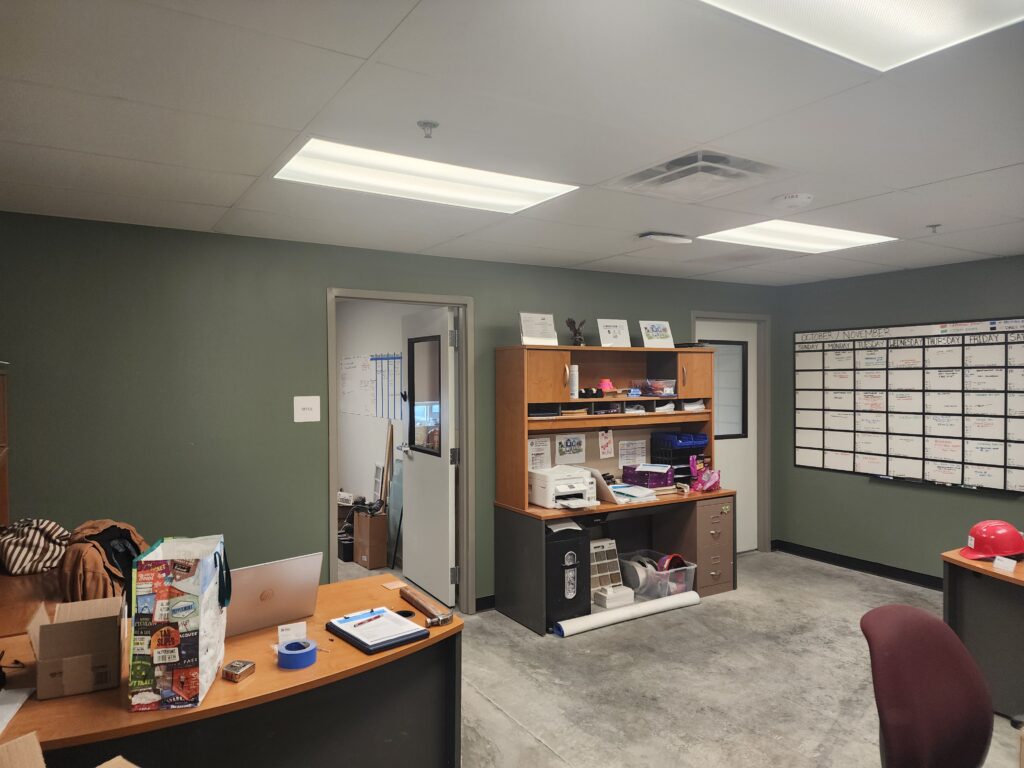 Interior view of an industrial facility office workspace with desks, storage cabinets, planning boards, and modern lighting