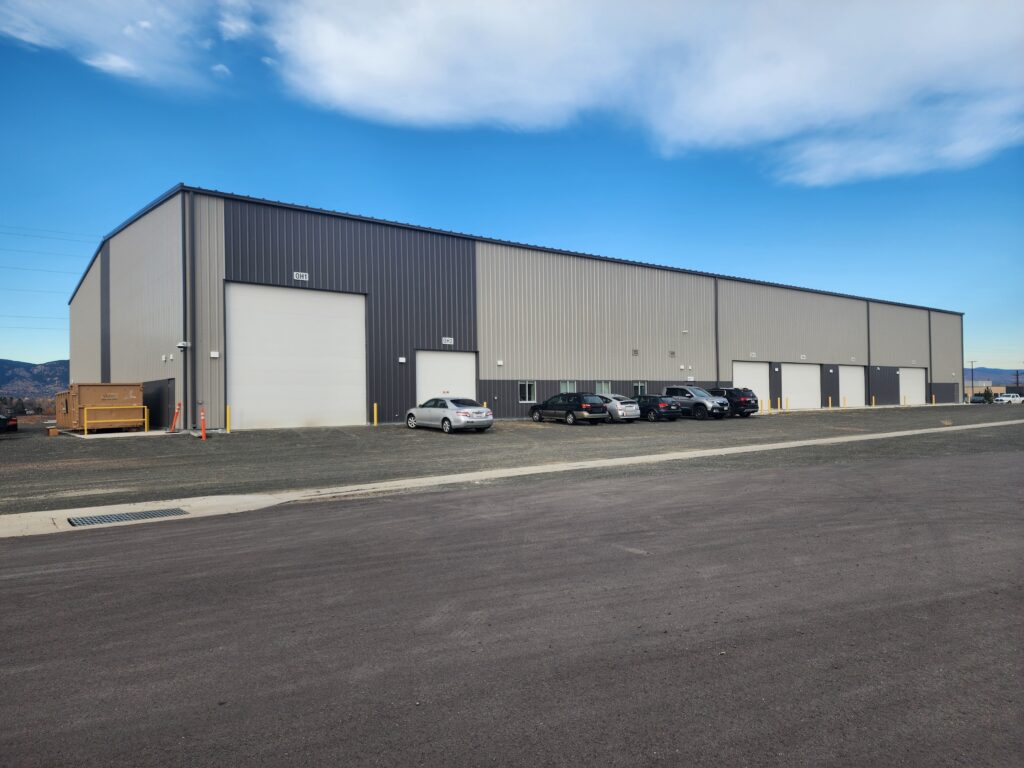 Large modern industrial warehouse building with metal exterior walls, loading doors, parking area, and mountain landscape background