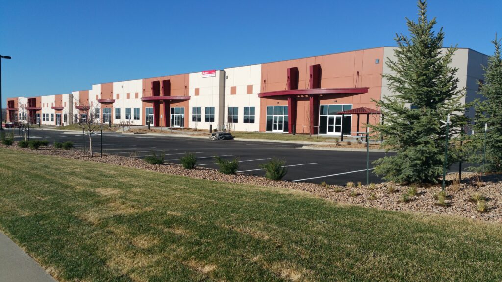 Large modern industrial facility exterior with multiple entrances, glass windows, parking area, and landscaped surroundings