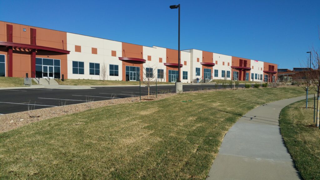 Modern industrial building exterior with glass windows, landscaped lawn, sidewalk, and parking area