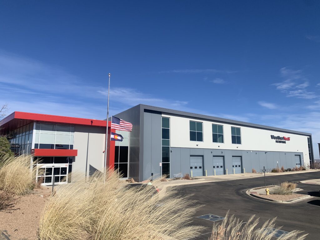 Modern industrial facility with gray and red exterior design, large warehouse doors, glass entrance, and landscaped surroundings
