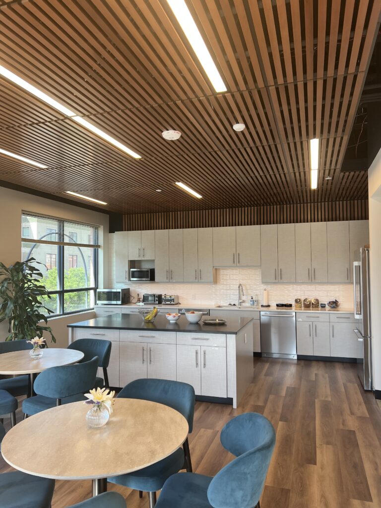 Modern office breakroom kitchen with white cabinets, island counter, dining tables, blue chairs, and wooden ceiling design