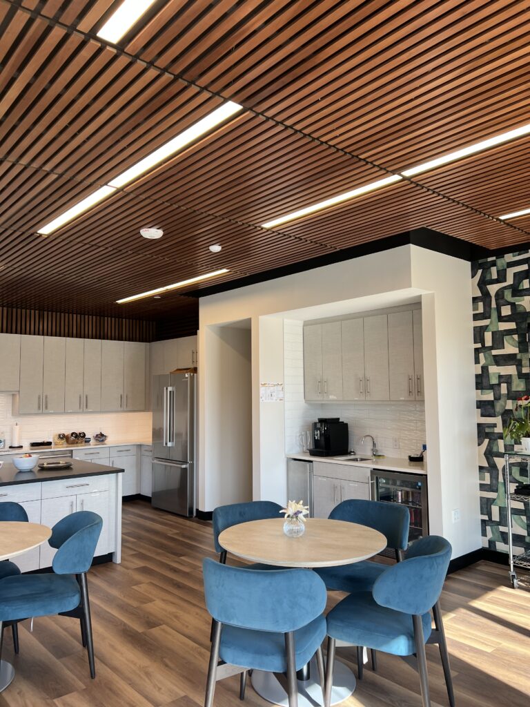 Modern office kitchen with white cabinets, dining area, blue chairs, wooden ceiling, and contemporary workplace interior design