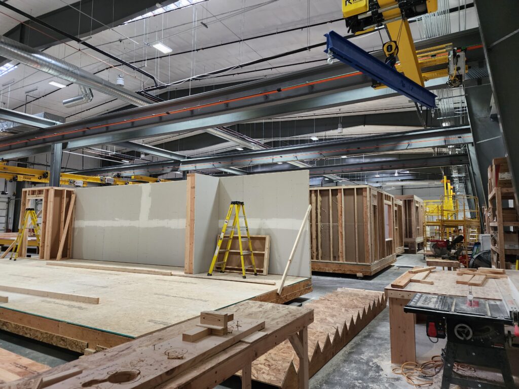 Modular construction facility interior with prefabricated wall panels, timber framing, overhead cranes, and assembly workstations
