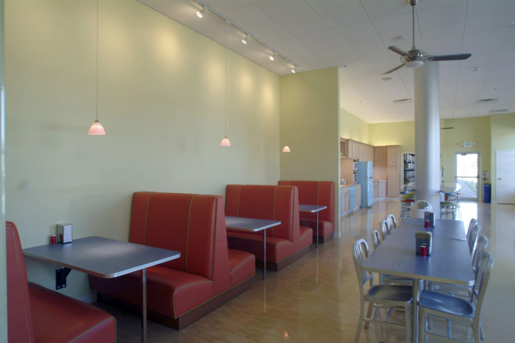 Modern office cafeteria with booth seating, dining tables, kitchenette area, and contemporary workplace interior design