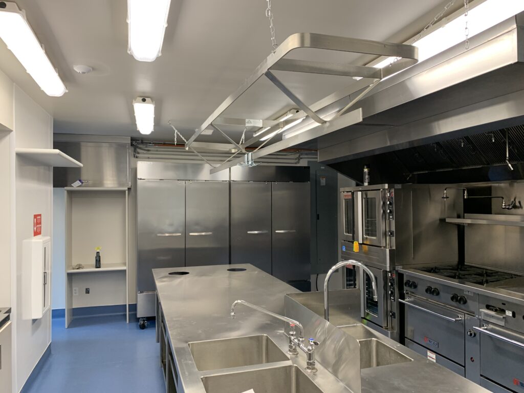 Commercial kitchen with stainless steel preparation tables, industrial ovens, ventilation hood, and storage equipment