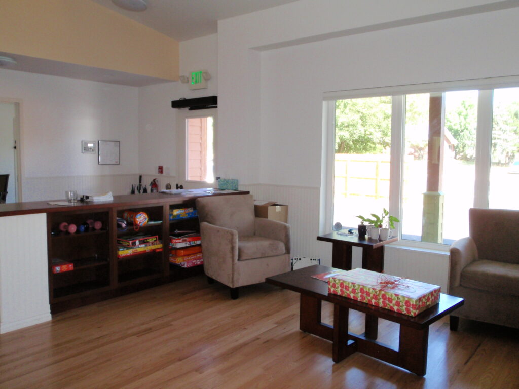Community facility lounge interior with seating chairs, coffee table, board game storage, large windows, and welcoming design