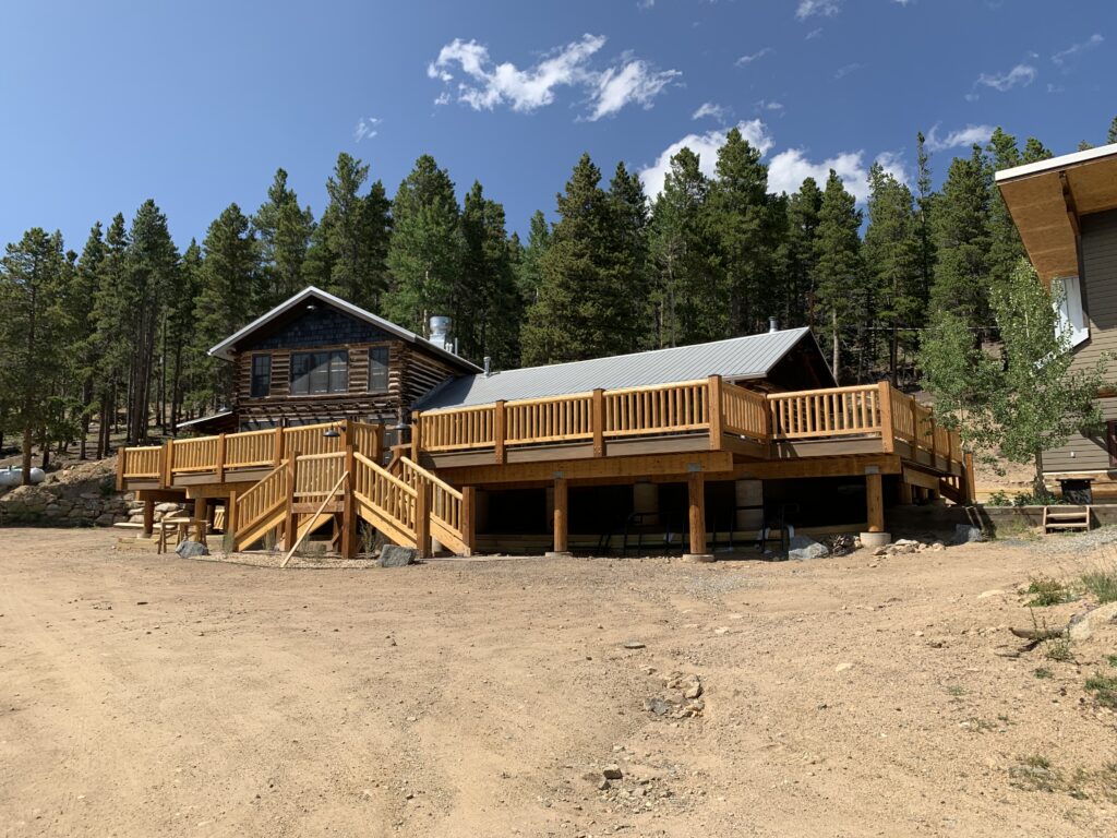 Large elevated wooden deck with stairs and railings attached to a mountain cabin surrounded by pine trees