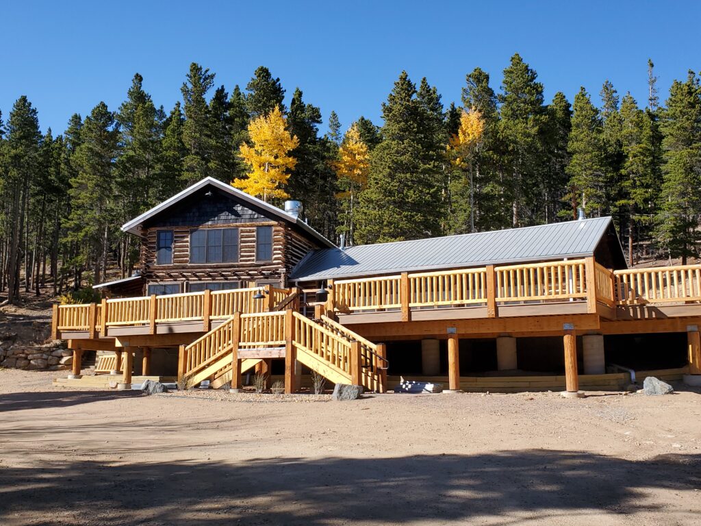 Rustic mountain cabin with a large elevated wooden deck, timber railings, and outdoor living space surrounded by pine trees