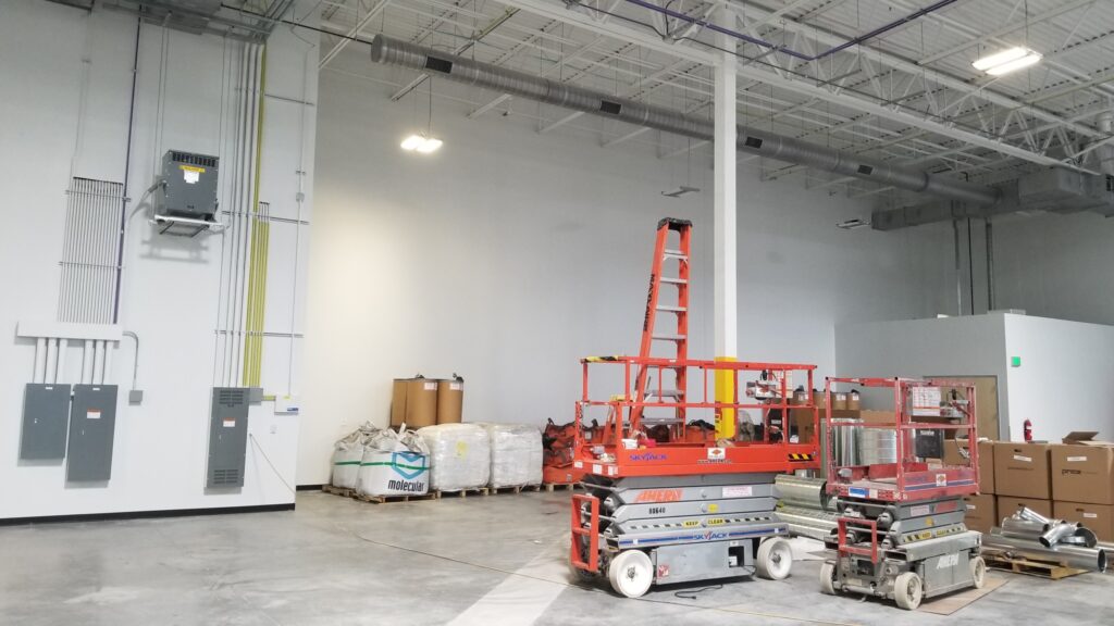 Large industrial warehouse interior with construction equipment, HVAC systems, electrical panels, and installation work in progress