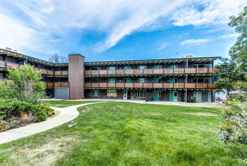 Modern multi-story apartment building with balconies, brick exterior, green lawn, and landscaped residential surroundings