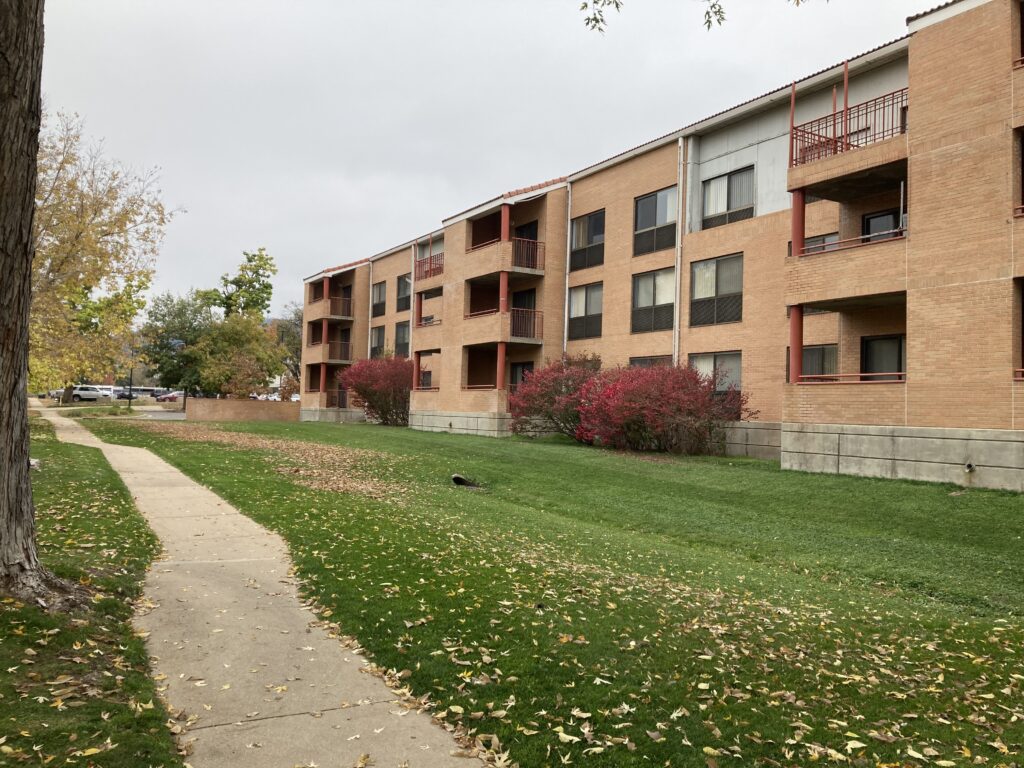 Multi-story brick apartment building with balconies, large windows, green lawn, and pedestrian walkway