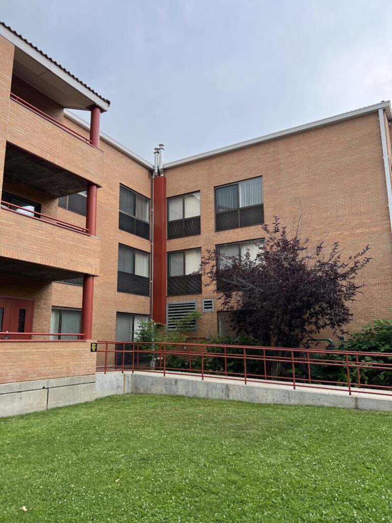 Exterior view of a modern brick building with large windows, red accents, balconies, and landscaped lawn area