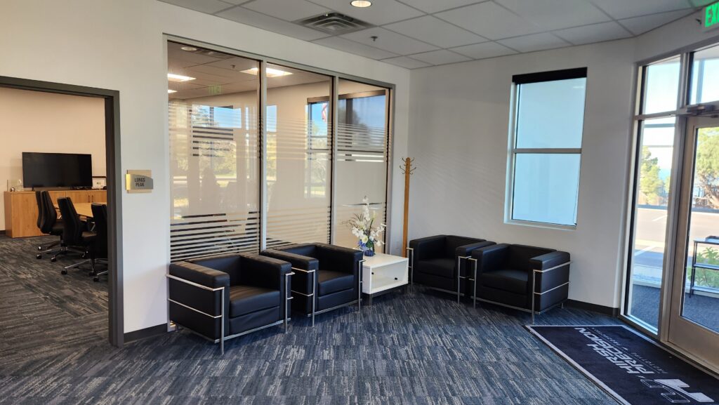 Modern office waiting area with black lounge chairs, glass partition offices, carpet flooring, and natural lighting