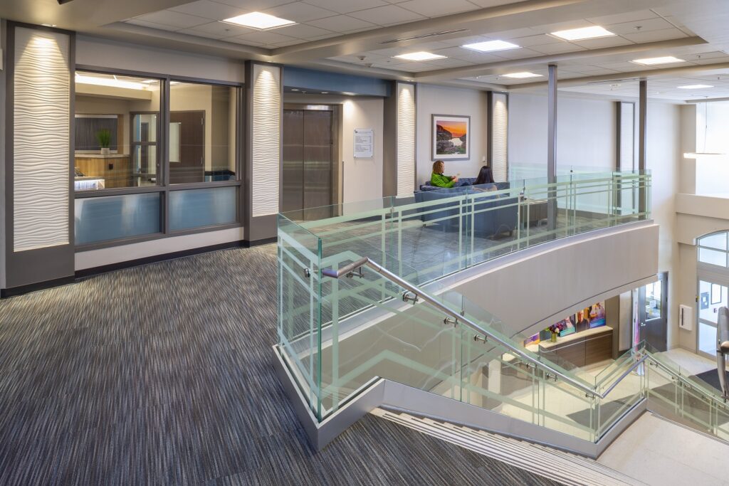 Modern healthcare facility interior featuring glass staircase, corridor, patient seating area, elevator lobby, and contemporary design elements