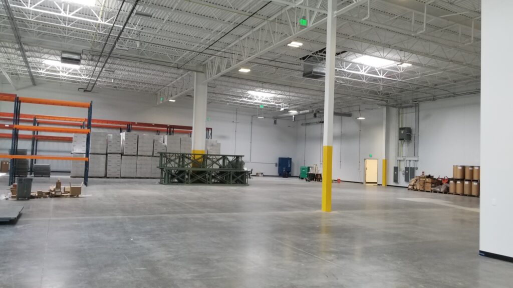 Large industrial warehouse interior featuring high ceilings, concrete flooring, storage racks, and open workspace area