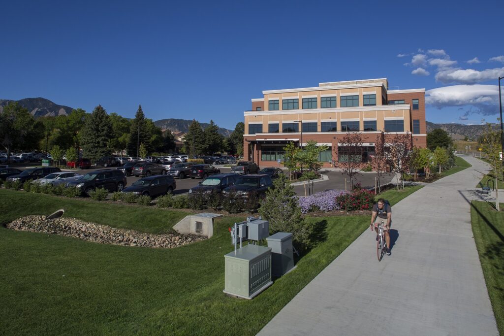 Modern university campus building with glass windows, landscaped grounds, parking area, and mountain background