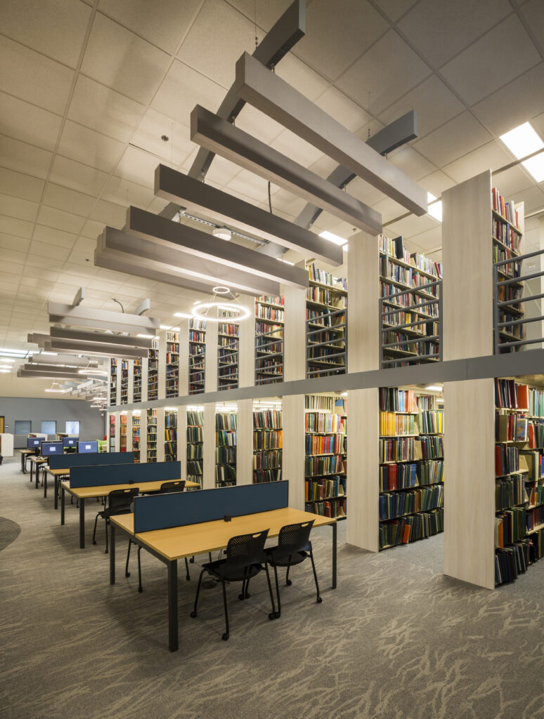 Modern university library interior with tall bookshelves, study desks, computer stations, and contemporary ceiling lighting