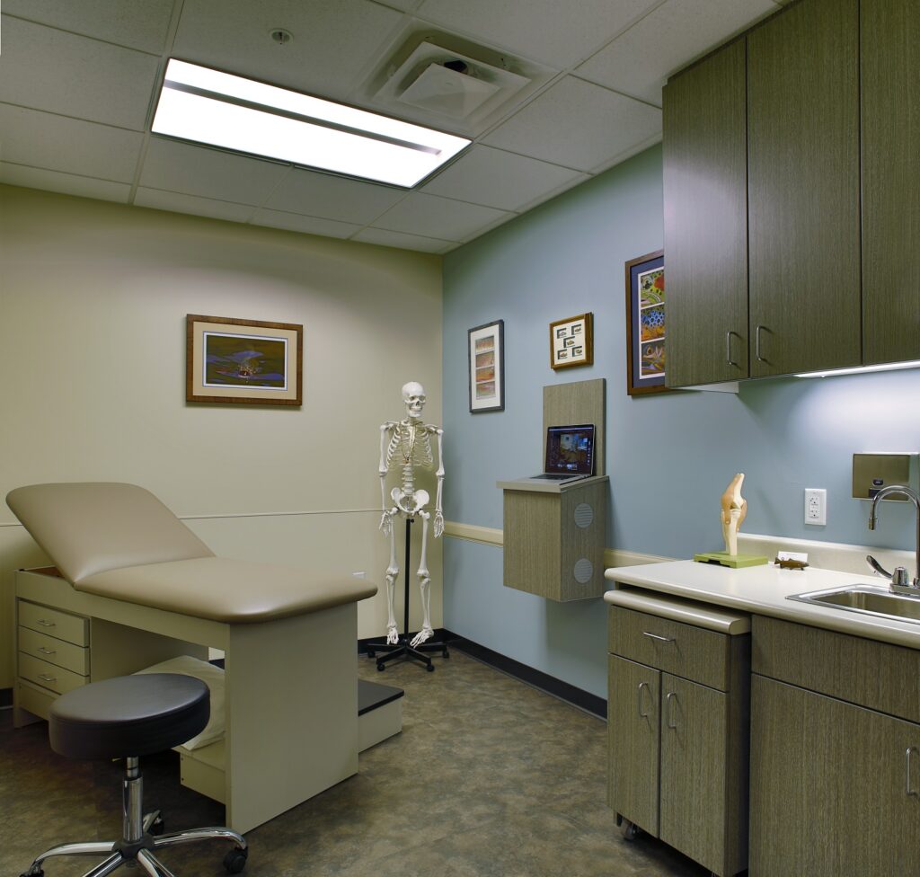 Modern orthopedic clinic examination room with patient table, skeleton model, medical cabinets, sink, and healthcare equipment