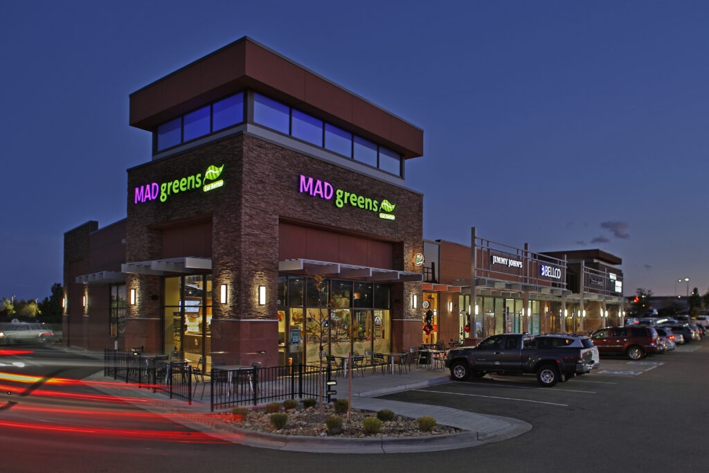 Modern retail center exterior at night featuring illuminated storefronts, restaurants, parking area, and contemporary commercial architecture